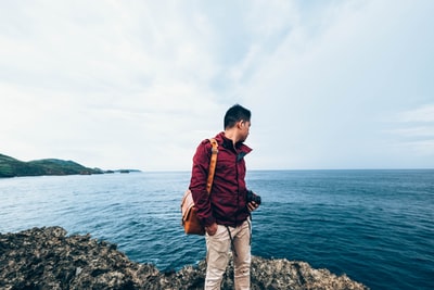 男人穿红色拉上拉链夹克站在岩石悬崖旁边的水域的白天
